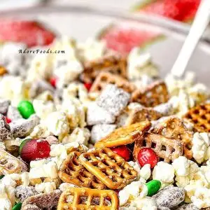 Festive homemade Chex Mix served in a bright red ceramic bowl for Christmas. The snack mix features golden brown toasted cereal squares, pretzel twists, round cheese crackers, and roasted nuts, all lightly coated with seasoning for a shiny, crunchy texture. The bold red color of the bowl adds a holiday touch, complementing the mix&rsquo;s warm tones of gold and brown, while the background hints at a cozy Christmas setting perfect for parties, gifting, or family snacking.