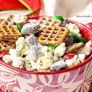 Festive homemade Chex Mix served in a bright red ceramic bowl for Christmas. The snack mix features golden brown toasted cereal squares, pretzel twists, round cheese crackers, and roasted nuts, all lightly coated with seasoning for a shiny, crunchy texture. The bold red color of the bowl adds a holiday touch, complementing the mix&rsquo;s warm tones of gold and brown, while the background hints at a cozy Christmas setting perfect for parties, gifting, or family snacking.