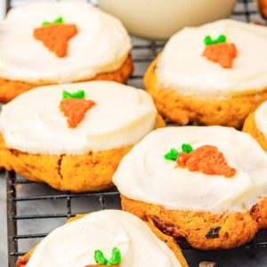 Close-up of soft and moist carrot cake cookies topped with creamy white cream cheese frosting, each decorated with a small carrot-shaped icing. The cookies are cooling on a black wire rack, with visible pieces of walnuts and carrot bits in the cookies. Perfect homemade carrot cake cookies with rich cream cheese icing, ideal for a delicious dessert or snack
