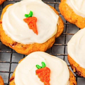Close-up of soft and moist carrot cake cookies topped with creamy white cream cheese frosting, each decorated with a small carrot-shaped icing. The cookies are cooling on a black wire rack, with visible pieces of walnuts and carrot bits in the cookies. Perfect homemade carrot cake cookies with rich cream cheese icing, ideal for a delicious dessert or snack