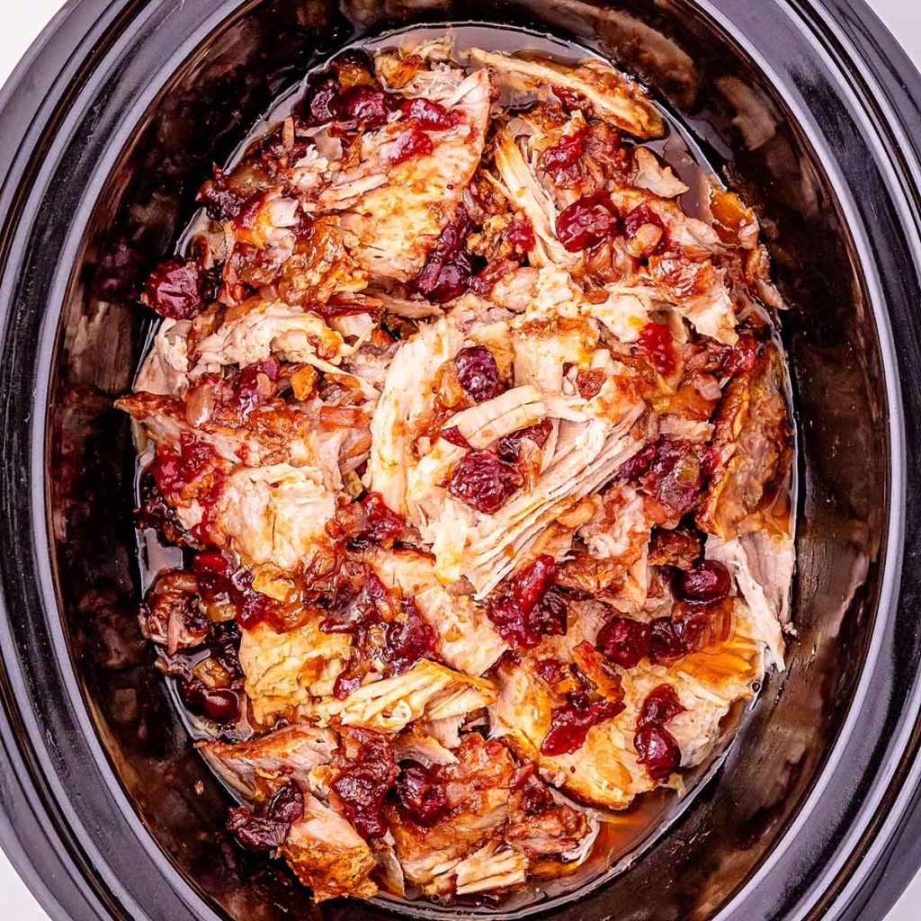 Close-up of a juicy cranberry pork loin resting in a crock pot, cooked low and slow until fork-tender. The pork is coated in a glossy cranberry glaze that clings to the meat, with whole cranberries nestled around the roast. The deep red sauce contrasts with the golden-brown seared edges of the pork, and steam rises gently from the pot, highlighting the rich, sweet-tart aroma of the holiday dish.