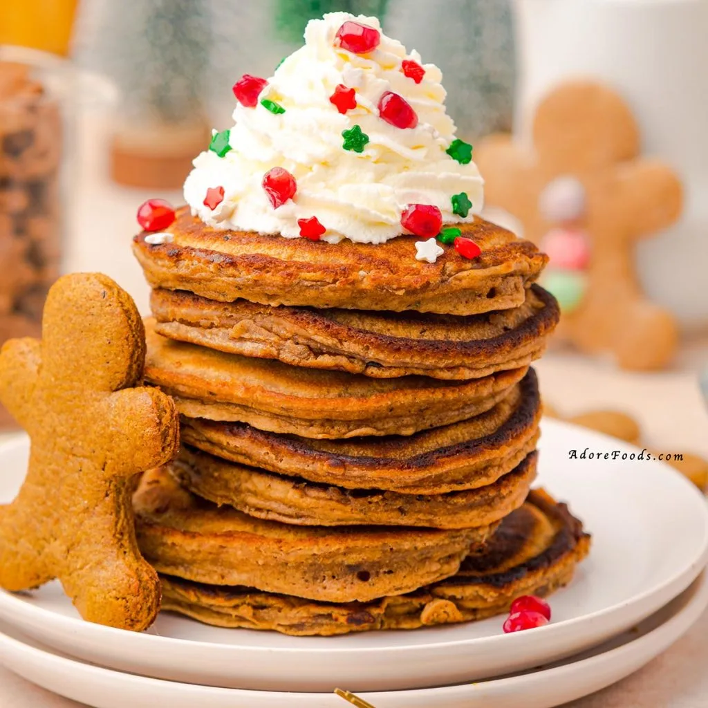 A tall stack of golden-brown gingerbread pancakes topped with a generous swirl of whipped cream, drizzled with glossy maple syrup, lightly dusted with powdered sugar, sprinkled with colorful festive sprinkles, and finished with bright red pomegranate seeds for a holiday-inspired breakfast presentation.