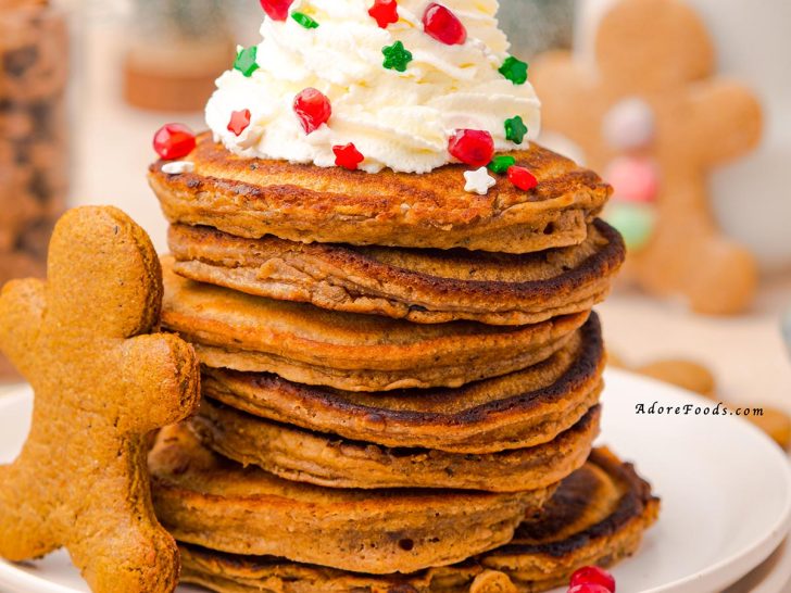 A tall stack of golden-brown gingerbread pancakes topped with a generous swirl of whipped cream, drizzled with glossy maple syrup, lightly dusted with powdered sugar, sprinkled with colorful festive sprinkles, and finished with bright red pomegranate seeds for a holiday-inspired breakfast presentation.