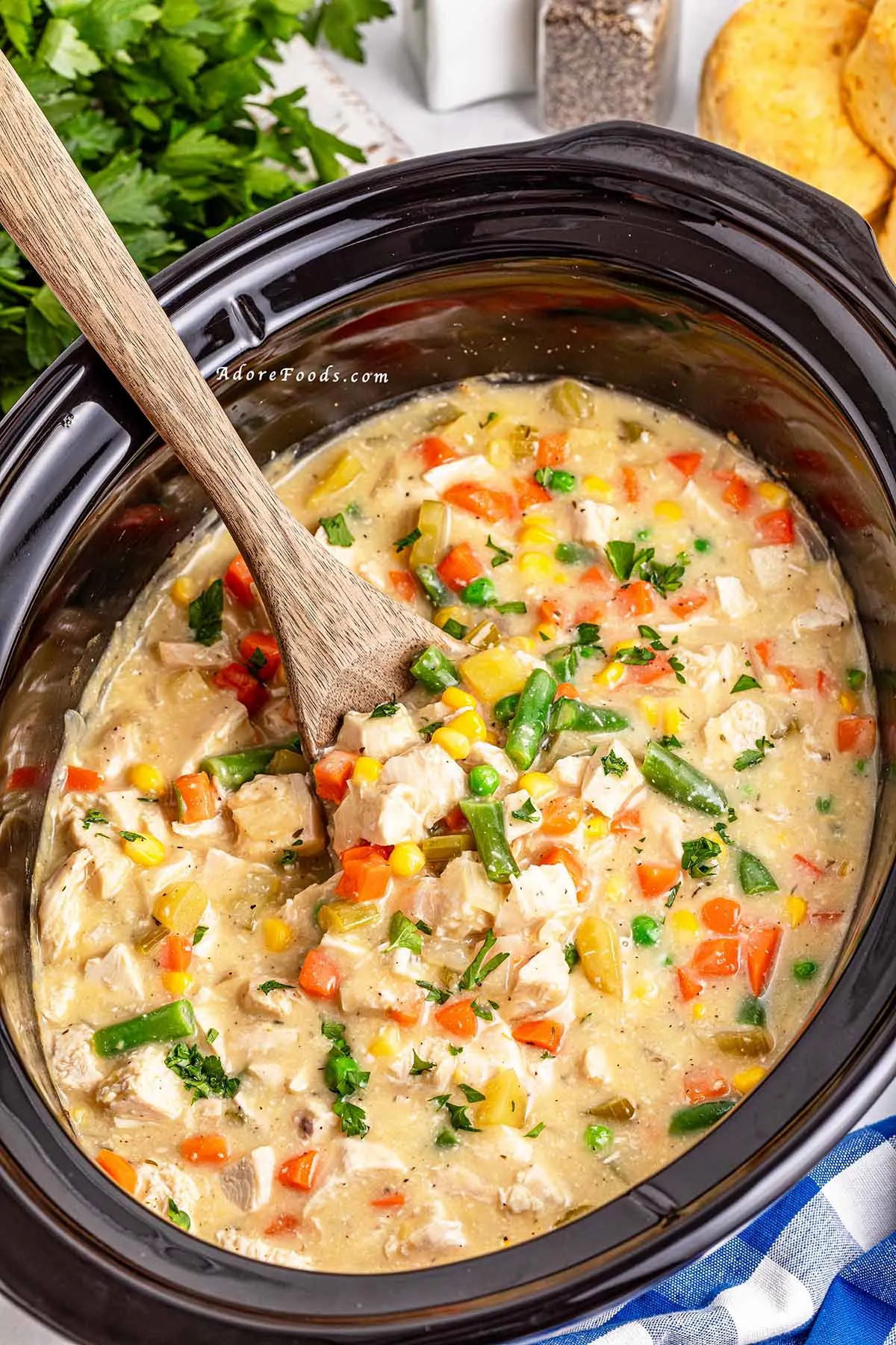 Overhead view of a creamy chicken pot pie filling cooked in a slow cooker, loaded with shredded chicken, tender potatoes, carrots, peas, and corn, served alongside golden-brown buttermilk biscuits on a plate, showcasing a hearty and comforting homemade crockpot dinner.