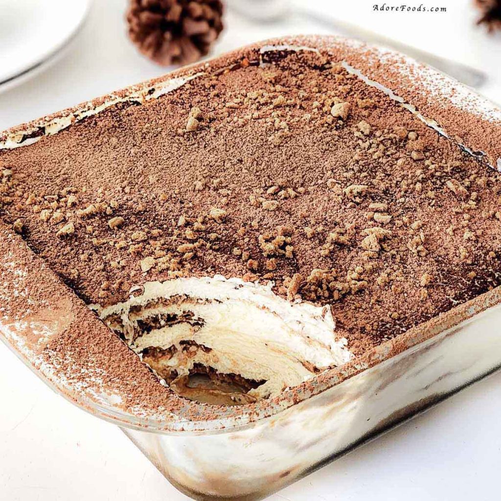 Homemade gingerbread tiramisu in a clear Pyrex baking dish, showing visible layers of dipped gingerbread cookies and creamy mascarpone eggnog filling, topped with a dusting of cocoa powder and crushed gingerbread crumbs, chilled and ready to serve as a festive Christmas dessert.