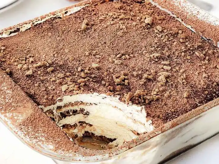 Homemade gingerbread tiramisu in a clear Pyrex baking dish, showing visible layers of dipped gingerbread cookies and creamy mascarpone eggnog filling, topped with a dusting of cocoa powder and crushed gingerbread crumbs, chilled and ready to serve as a festive Christmas dessert.