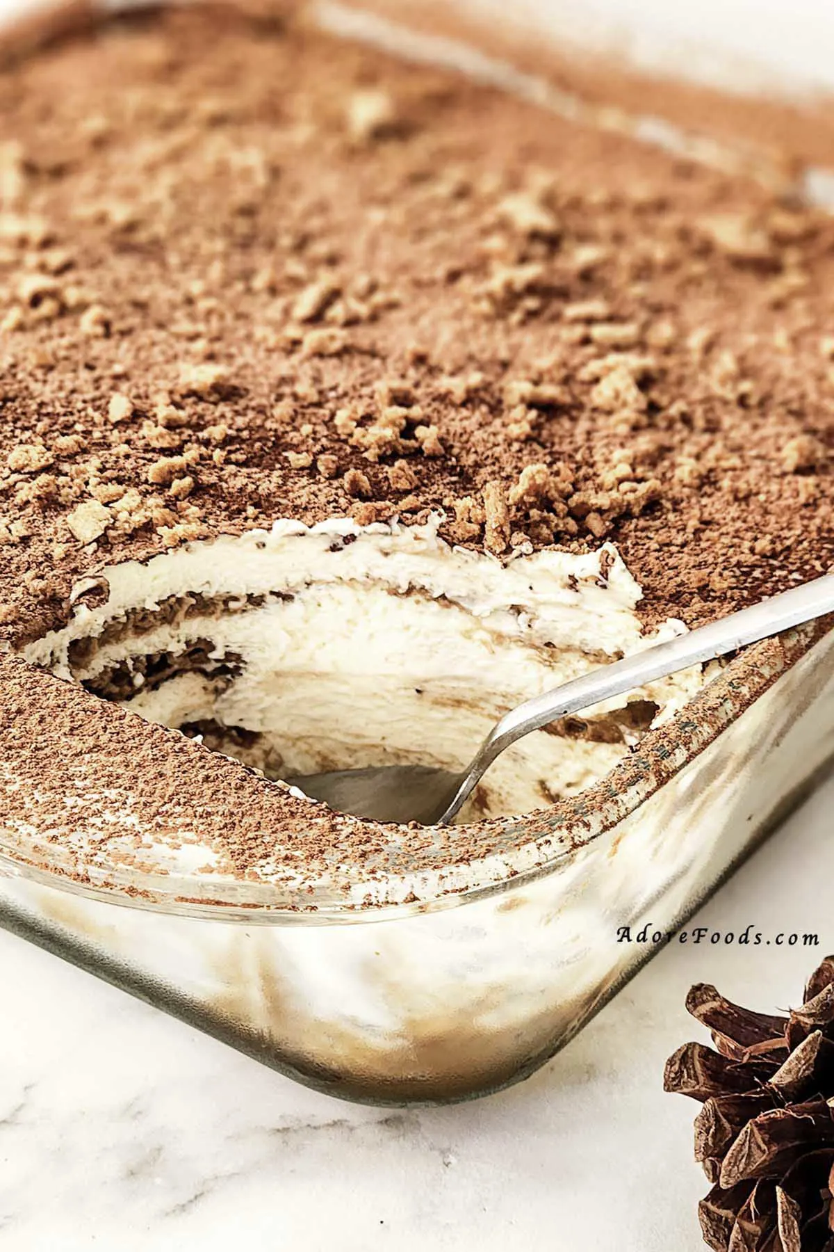 Homemade gingerbread tiramisu in a clear Pyrex baking dish, showing visible layers of dipped gingerbread cookies and creamy mascarpone eggnog filling, topped with a dusting of cocoa powder and crushed gingerbread crumbs, chilled and ready to serve as a festive Christmas dessert.