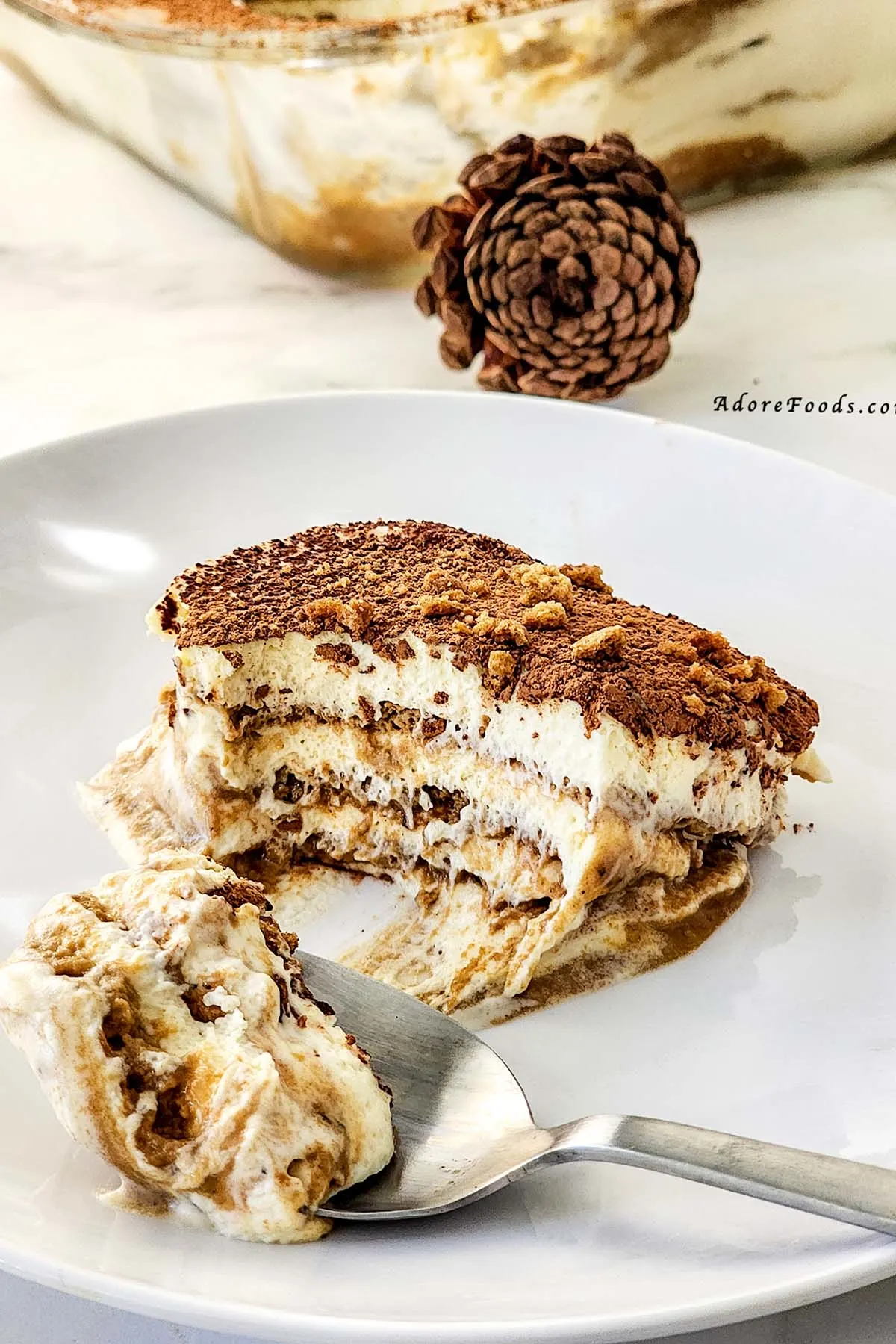 Slice of gingerbread tiramisu served on a white plate with a fork, showing distinct layers of spiced cookies, mascarpone cream, and cocoa topping, garnished with crushed gingerbread for a festive holiday dessert presentation.