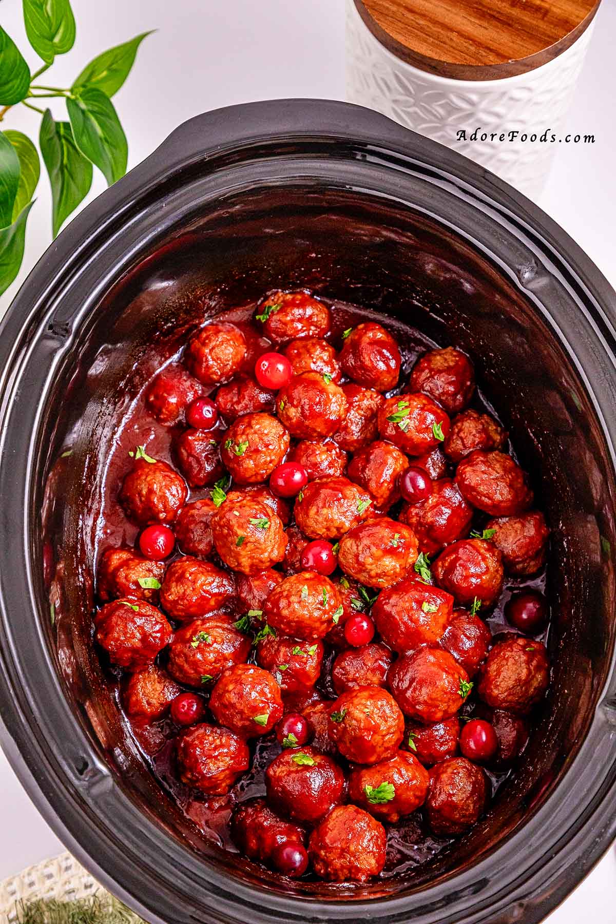 Overhead view of cranberry meatballs cooked in a black slow cooker, coated in a glossy sweet and tangy cranberry sauce and garnished with fresh herbs, ready to serve as an easy holiday appetizer.